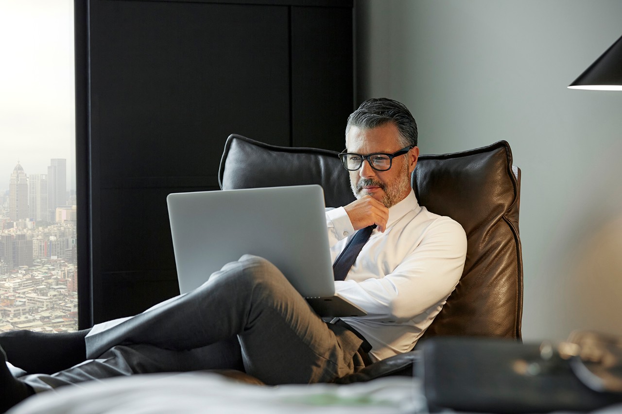 Full length of thoughtful businessman using laptop in hotel room. Professional is working. He is wearing eyeglasses.