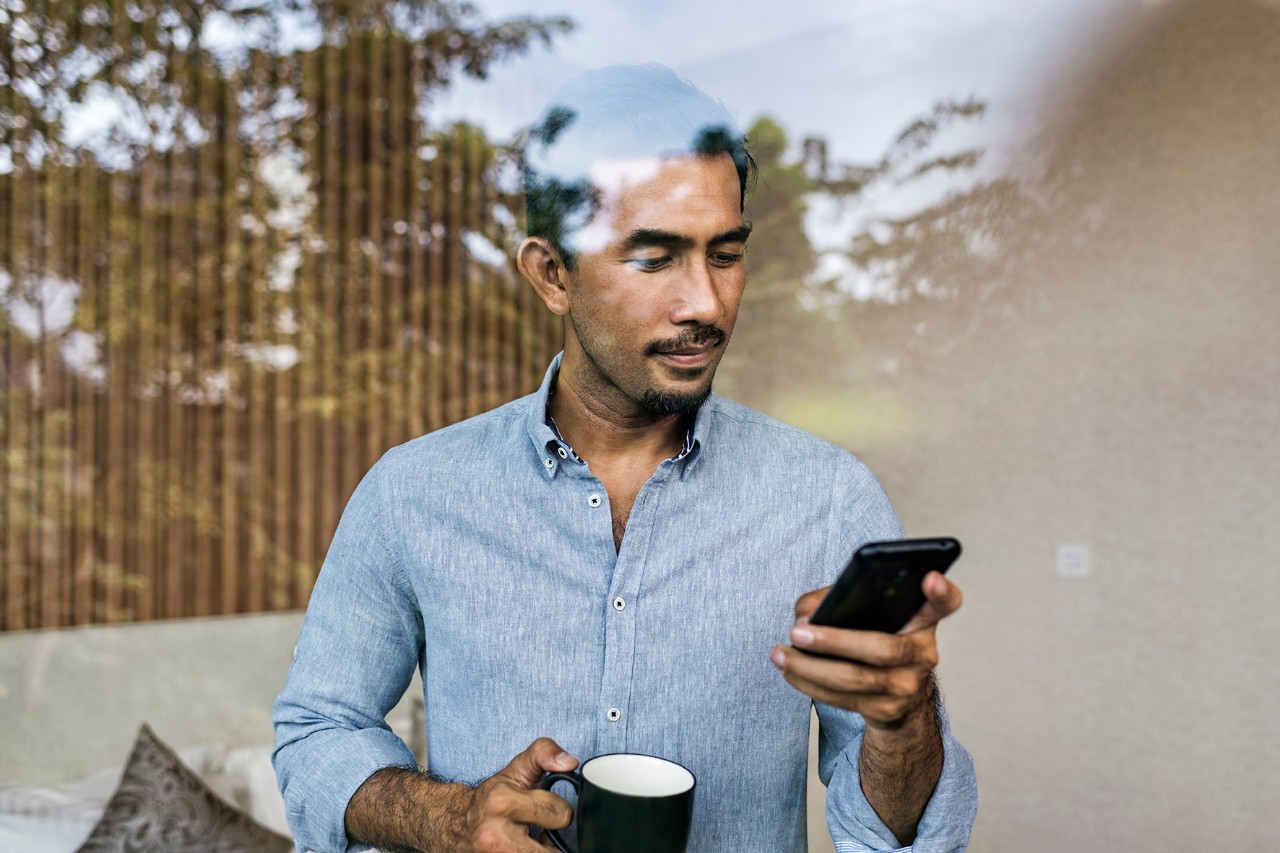 Un homme regardant son téléphone avec une tasse de café dans sa main derrière une vitre 